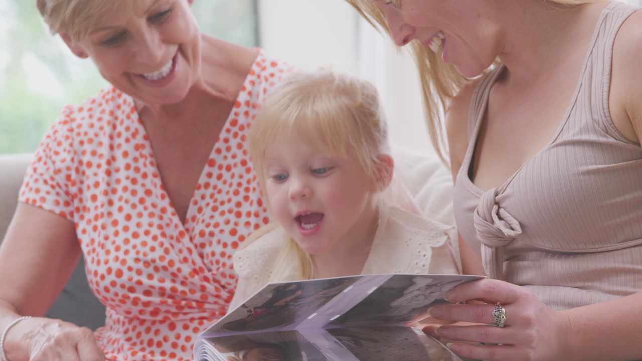 집 에서 어머니 와 할머니 와 함께 소파 에 앉아 함께 사진 앨범 을 보고 있는 손녀