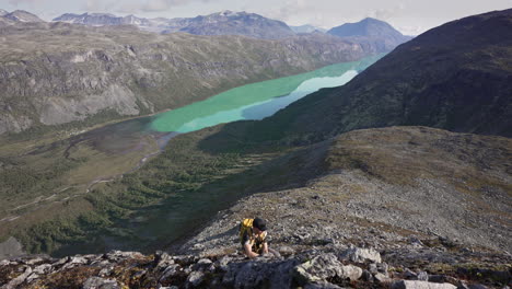 Wide-top-down-view-of-male-hiking-hill-side-with-green-lake-and-mountains-in-backgroud-in-early-sunny-autumn-morning