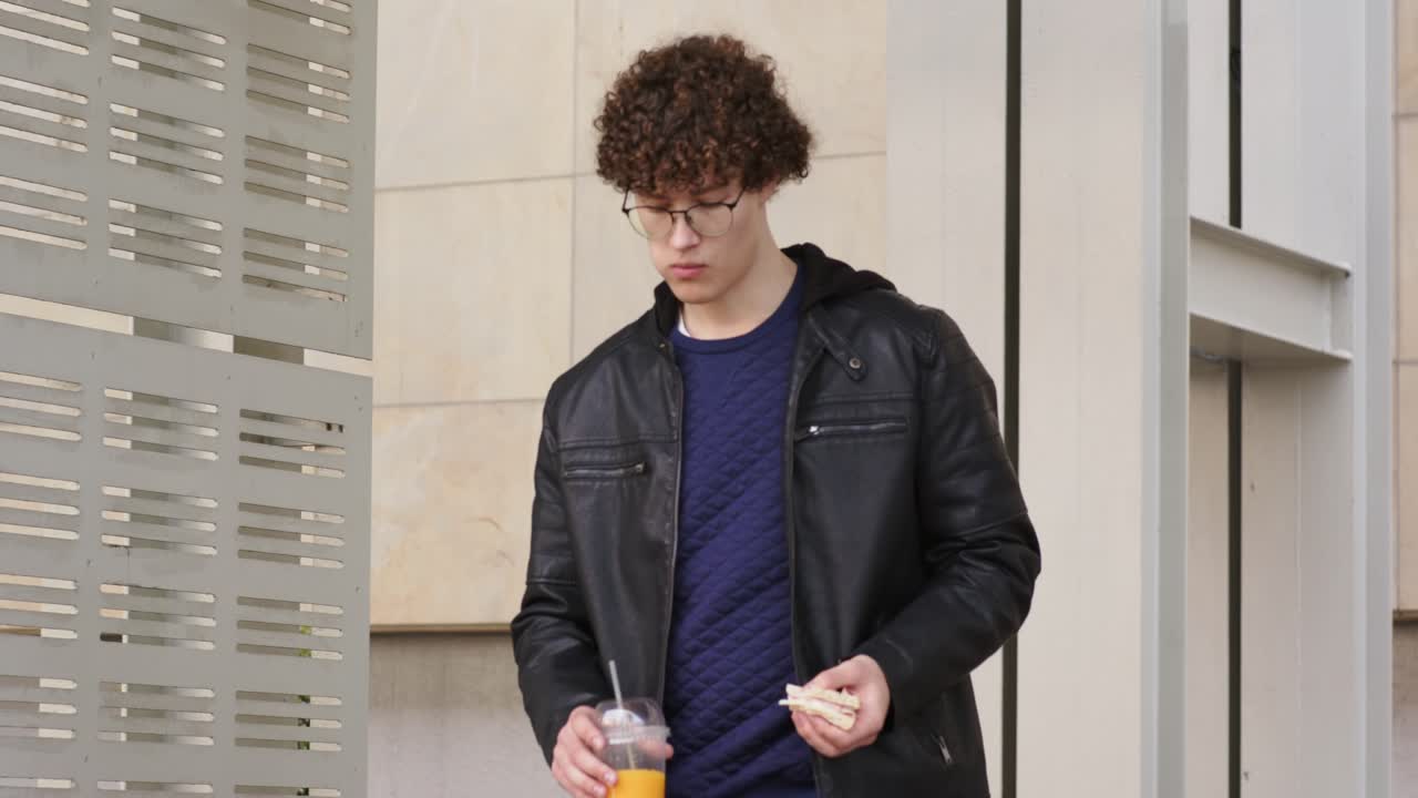 Homem na casa dos 20 anos caminhando em passarela pavimentada, mordendo sanduíche e bebendo suco enquanto verifica painel de metal.