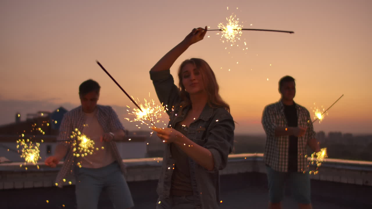 joven mujer en el techo mueve sus brazos y cuerpo hermosamente un baile con sus amigos en una noche de verano con gran luz bengal. su cabello sopla hermosamente en el viento.