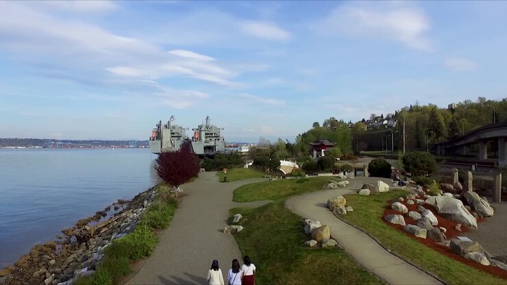 Women Walking At The Chinese Reconciliation Park In Tacoma, Washington With Two Cargo Ships Of The US Navy Reserve Fleet, Cape Island And Cape Intrepid, Docked On The Shore - aerial drone