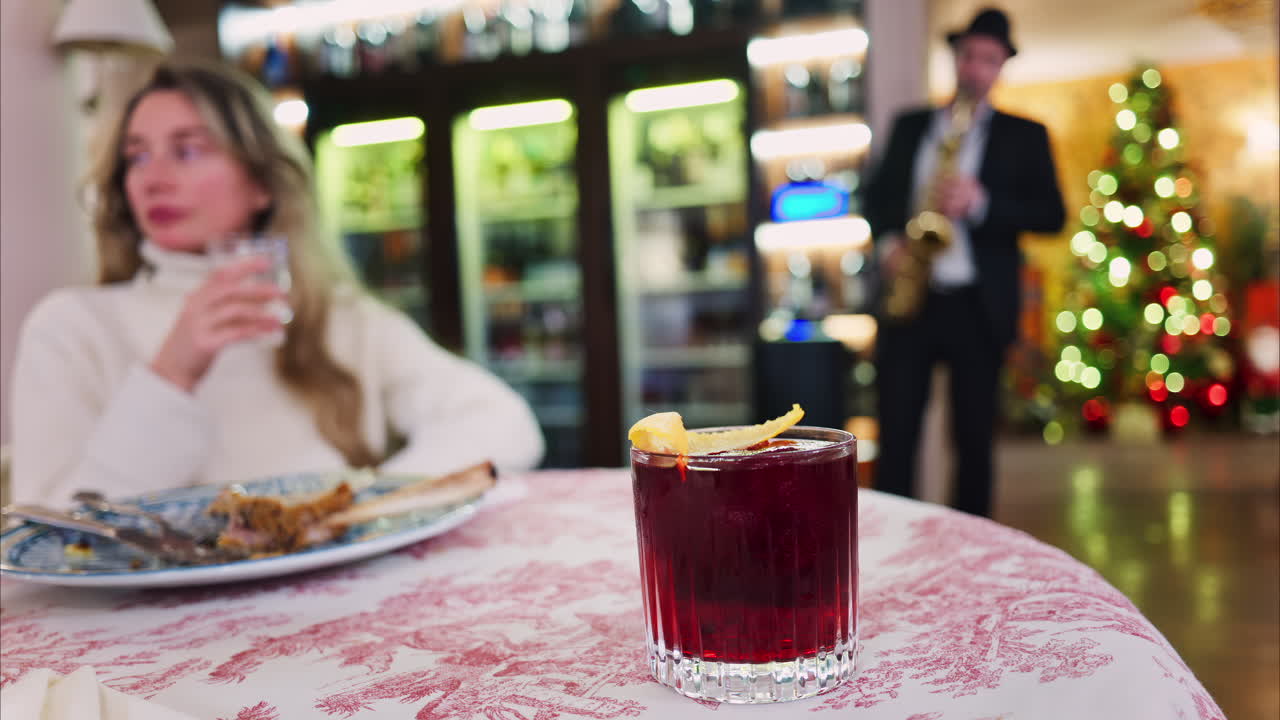 Close up of a negroni cocktail on a red and white tablecloth with a woman sitting at a restaurant on the background