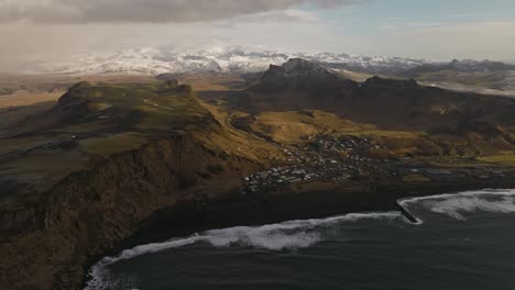 aerial footage showcasing the town of vik, iceland, with its dramatic coastal cliffs and snow-capped mountains in the background