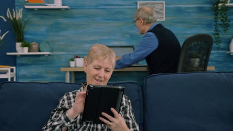 senior woman reading emails on digital tablet sitting on sofa at home