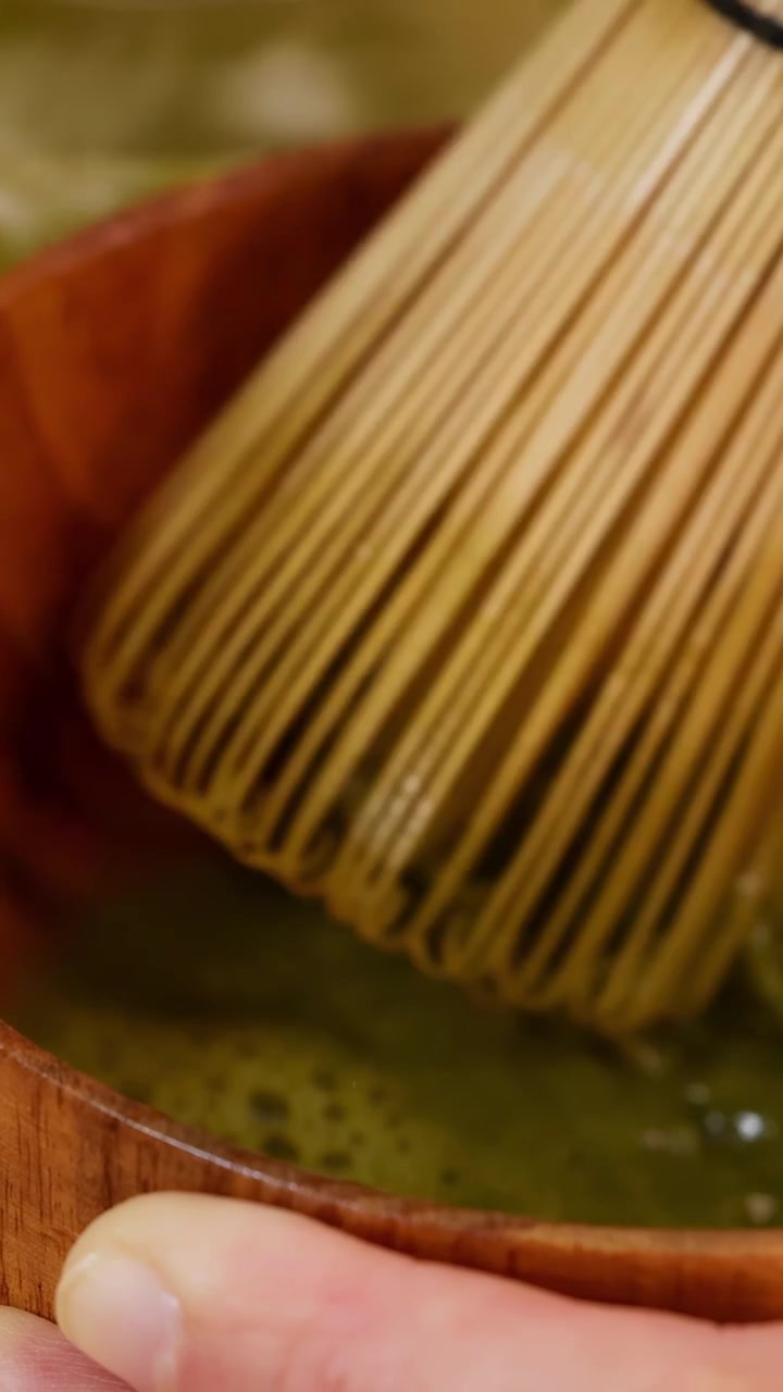 Primo piano di mani che frustano tè verde matcha in una ciotola di legno con un movimento fluido della telecamera.
