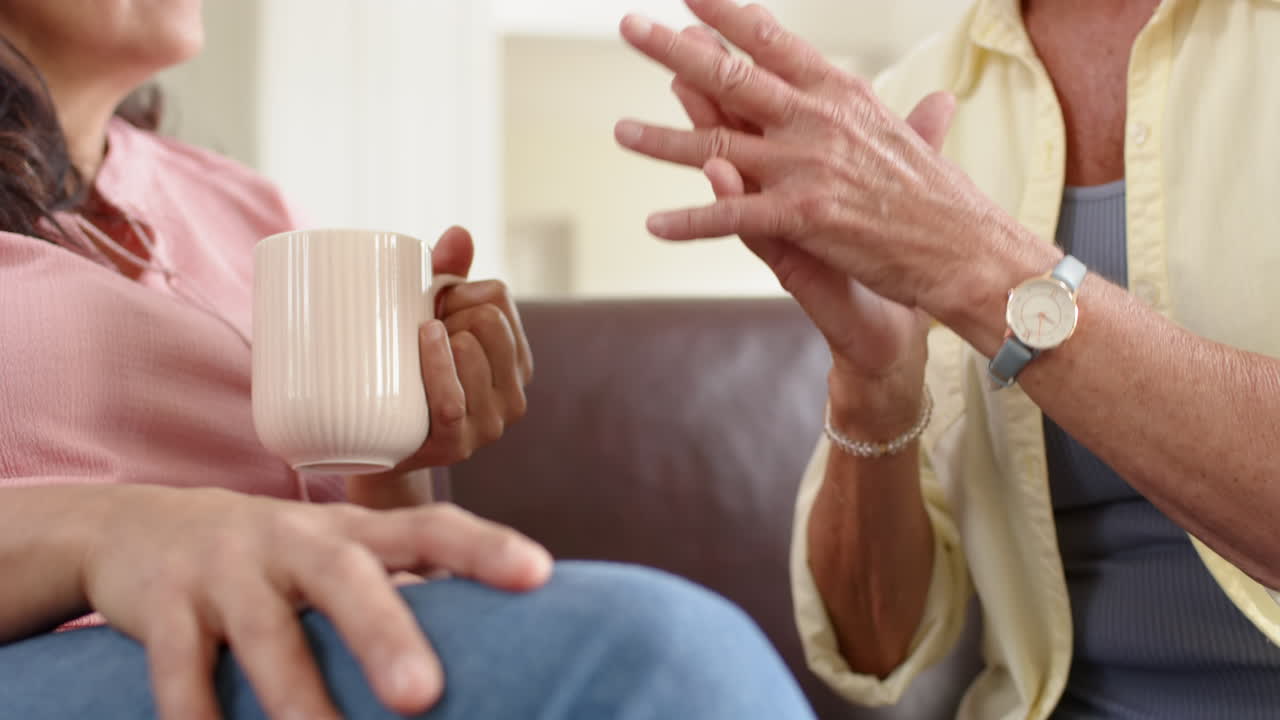mujeres mayores felices y diversas discutiendo y tomándose de la mano en el sofá en la sala de estar soleada, cámara lenta