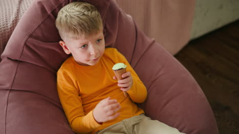 white child seated comfortably on pink beanbag indoors, holding ice cream cone and enjoying sweet treat, casually playing with leg, relaxed in cozy room with soft furnishings and wooden floor