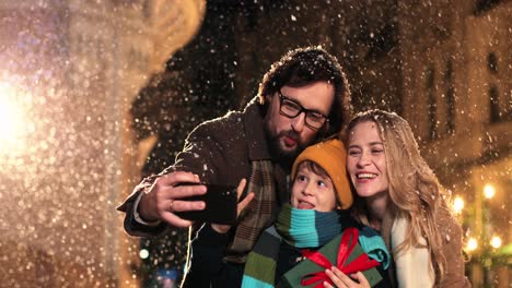 vista de cerca de una familia feliz caucásica sosteniendo regalos y haciéndose un selfie mientras nieva en navidad
