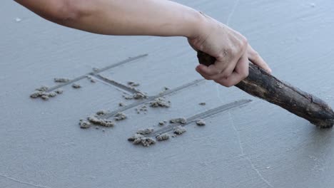 a hand uses a stick to draw letters on smooth, wet sand, creating textured impressions.