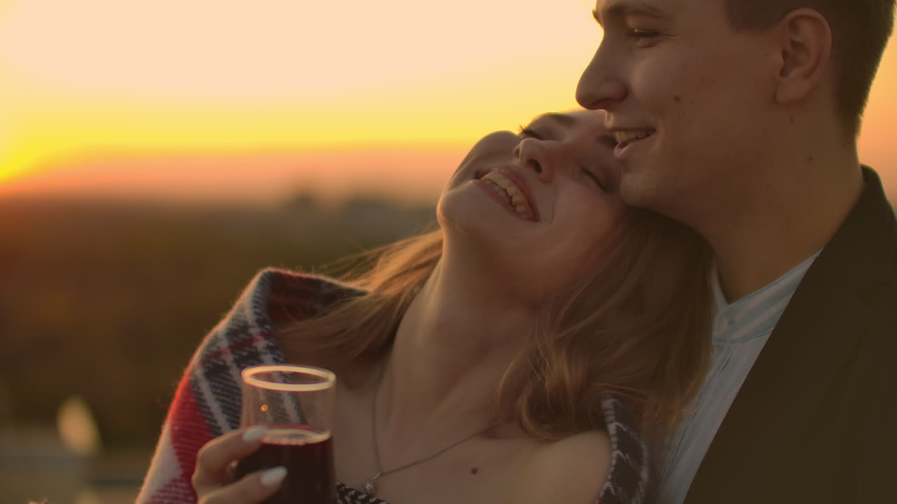 hombre y joven mujer bonita sentados en el sofá sosteniendo copas de vino y besándose en la terraza de la azotea al atardecer