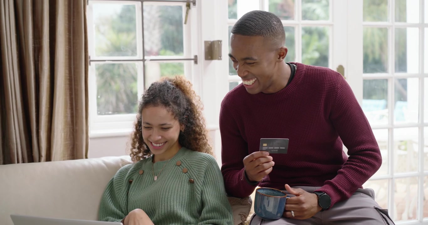 Couple sitting on sofa near window, man showing payment card while woman clicking laptop to buy