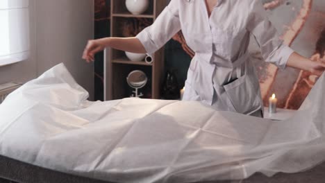 female massage therapist in a white robe covers the massage table with a disposable sheet