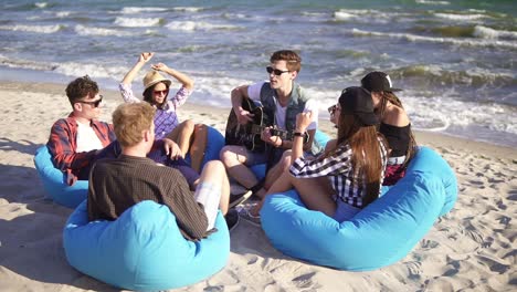 Young-man-playing-guitar-among-group-of-friends-sitting-on-easychairs-on-the-beach-and-singing-on-a-summer-evening.-Slowmotion-shot