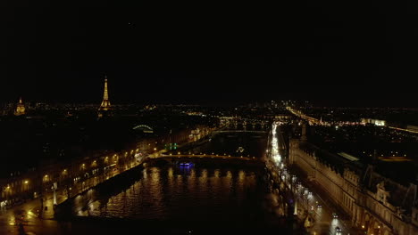 Forwards-fly-above-Seine-river-in-city-centre-at-night.-Busy-road-on-waterfront,-illuminated-buildings-and-landmarks.-Paris,-France