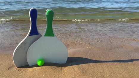 Beach-tennis-paddle-rackets-and-green-balls-on-sandy-beach-with-waves-breaking-on-shore