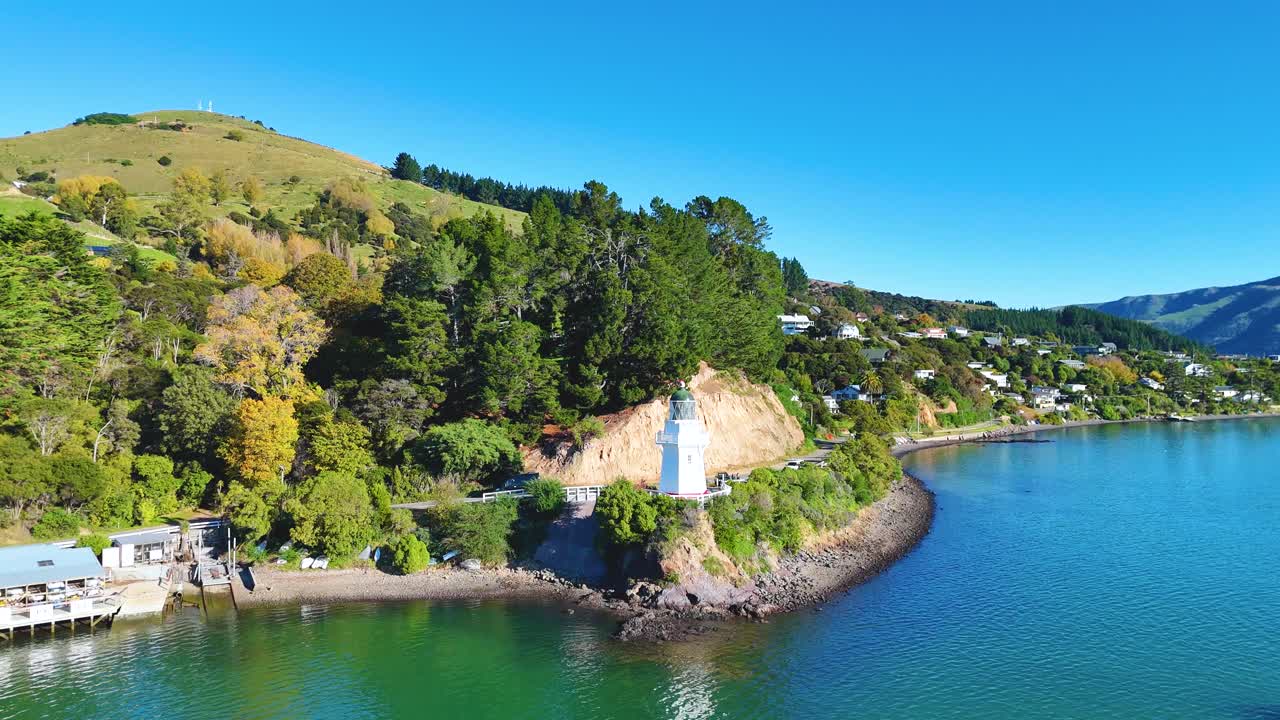 Aerial footage captures Akaroa's lighthouse, lush greenery, and tranquil harbor under clear blue skies, showcasing natural beauty and serene landscapes