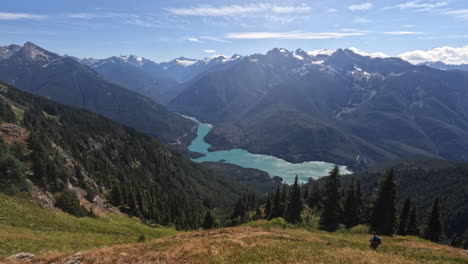 The-gorgeous-view-from-Sourdough-Mountain,-person-hiking-down-the-meadow-with-alpine-peaks-in-the-background