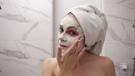 Portrait-of-young-pretty-woman-with-white-towel-on-head-is-applying-fresh-white-thick-mask-and-scrub-on-the-face.-Natural-organic-cosmetics-for-skin-care.-Woman-after-shower,-face-skin-care