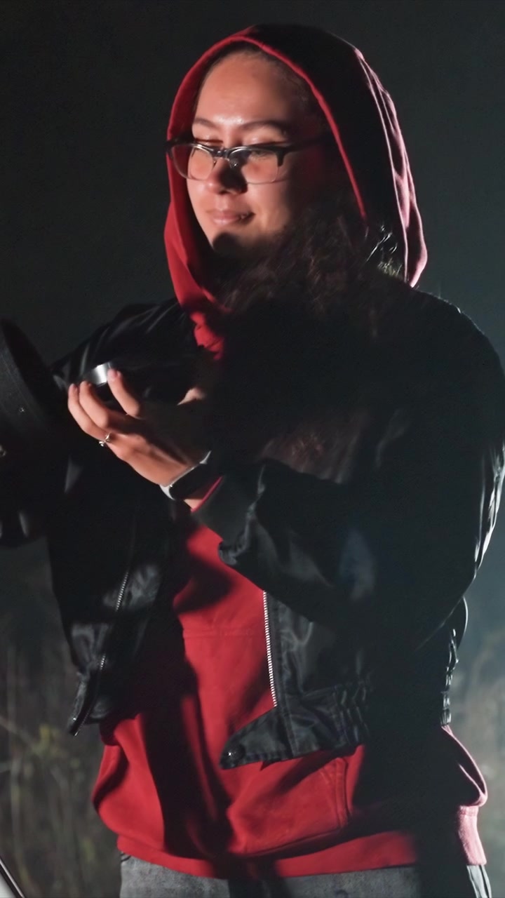 Subject wears black jacket and rings while handling device. Background shows faint tree outlines in darkness. Subject maintains contact near jacket zipper while adjusting