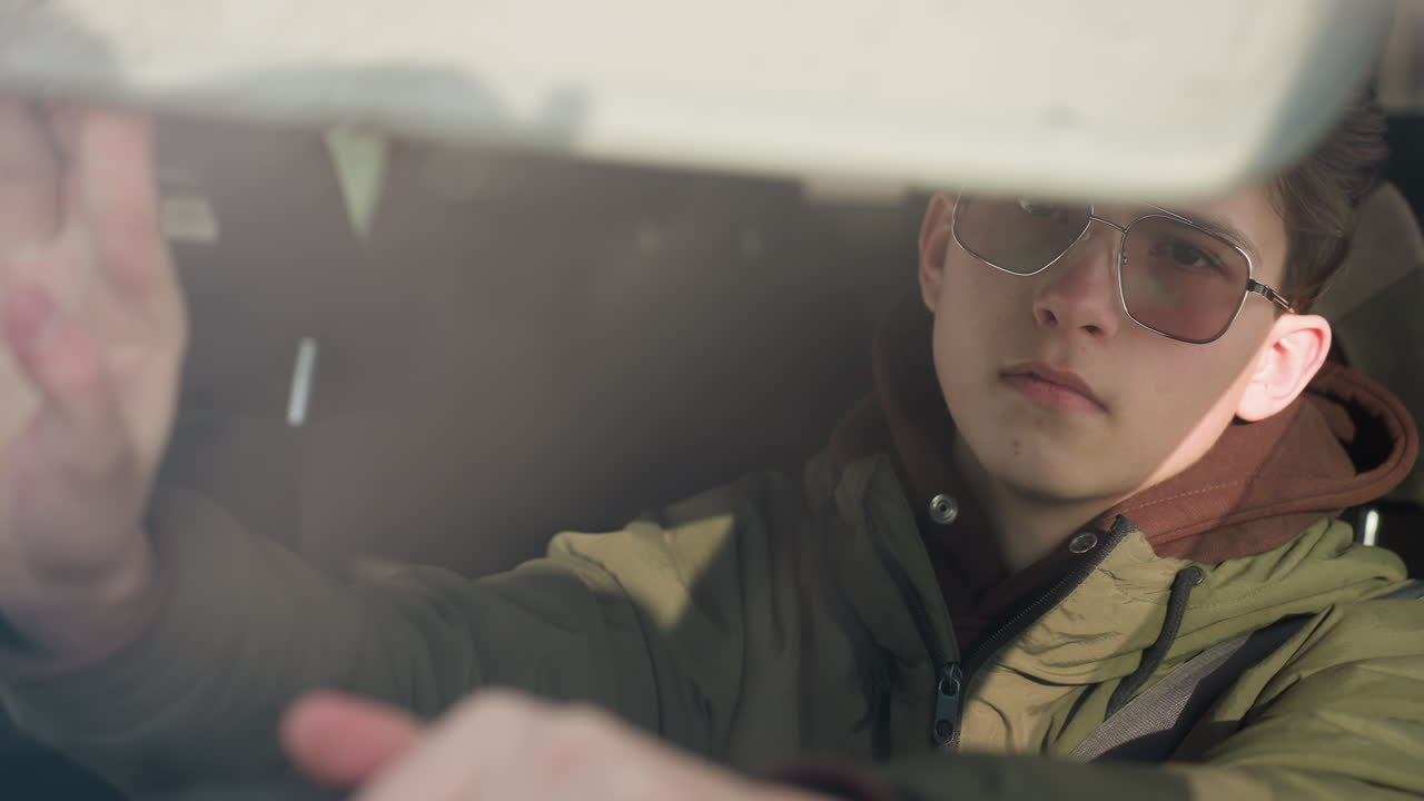 close up driver in winter jacket and hoodie wearing tinted glasses with sunlight reflections on face while leaning forward in car cabin, steady gaze through windshield, hand gripping steering wheel