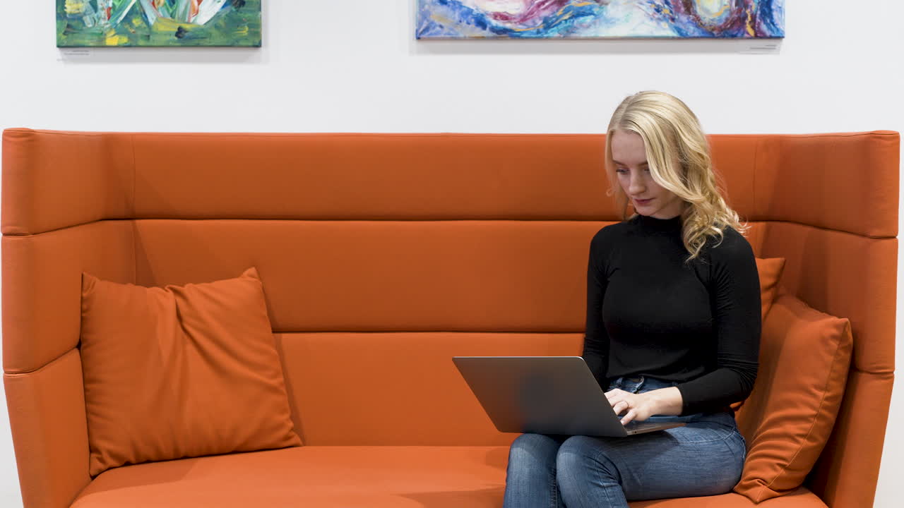 молодая женщина сидит на оранжевом офисном диване, работает на ноутбуке