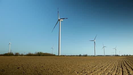 Wind-turbines-stand-tall-in-a-plowed-field-under-a-clear-blue-sky,-showcasing-renewable-energy