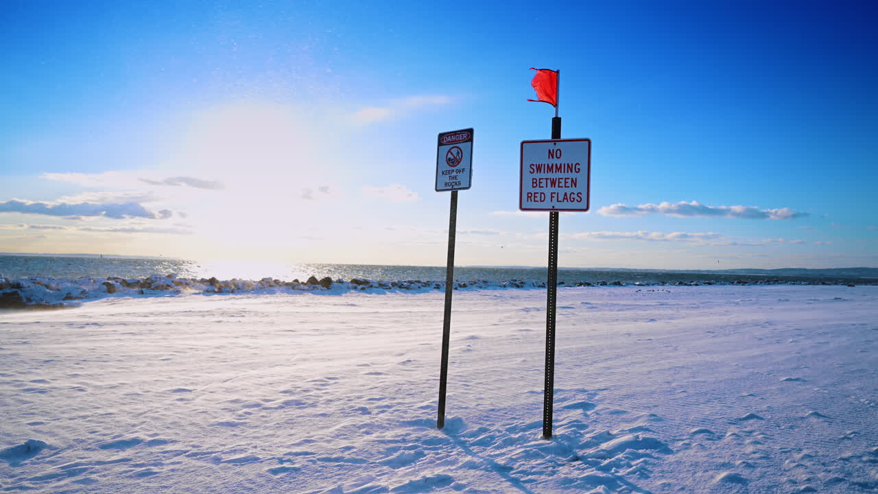 Segnali di avvertimento e bandiera rossa su una spiaggia invernale innevata. Segnali di divieto di balneazione e pericolo su una spiaggia innevata sotto la luce intensa del sole a New York City.