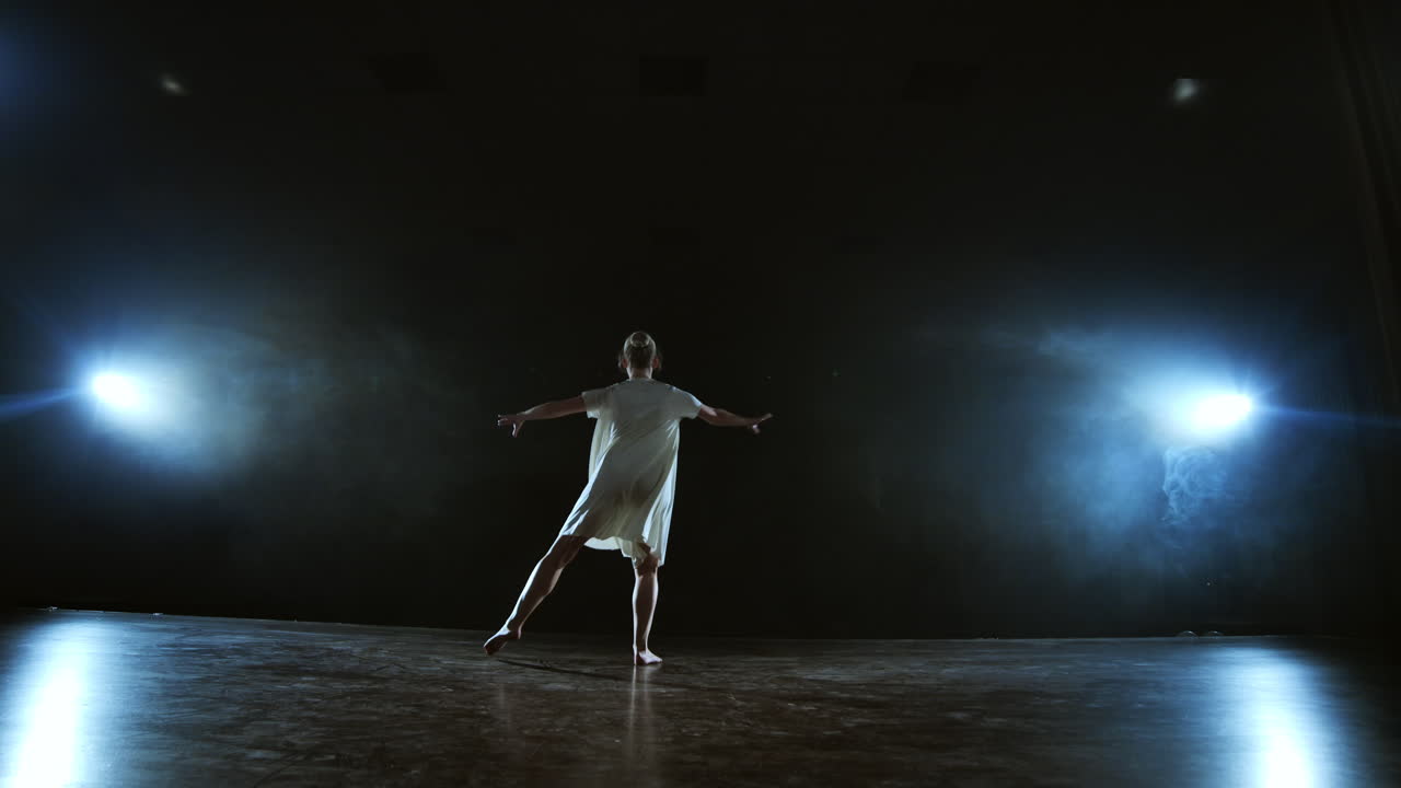 la telecamera zoom sposta la ballerina attraverso il palco con software e fumo. un moderno balletto drammatico una donna con un vestito bianco gira su una gamba e salta..