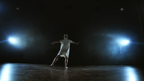 The-zoom-camera-moves-the-woman-dancer-across-the-stage-with-software-and-smoke.-A-modern-dramatic-ballet-a-woman-in-a-white-dress-spins-on-one-leg-and-jumps..