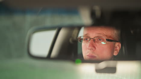 a close-up shot captures a man s face reflected in a car s rearview mirror, showing a focused and serious expression. the man is wearing glasses