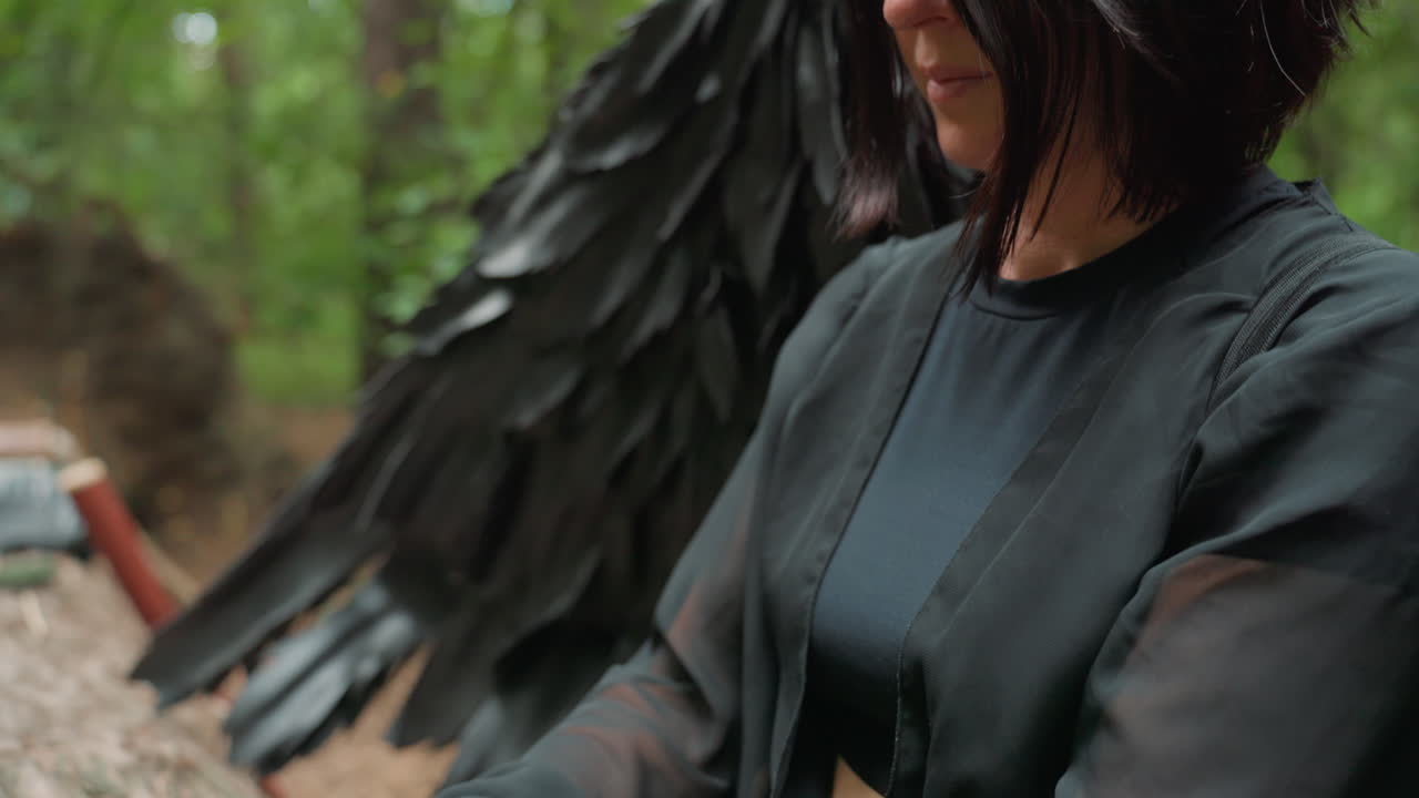 Side view of black fairy gently plucking metal tines on small green thumb piano placed on rough bark of fallen trunk in forest, soft light revealing flowing sleeves