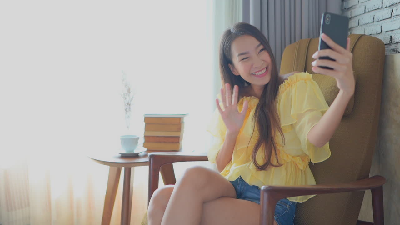 Smiling pleasant-looking Asian girl waves palm and says hello to the camera of a modern smartphone, makes a video call and smiling while sitting on the old armchair next to the window