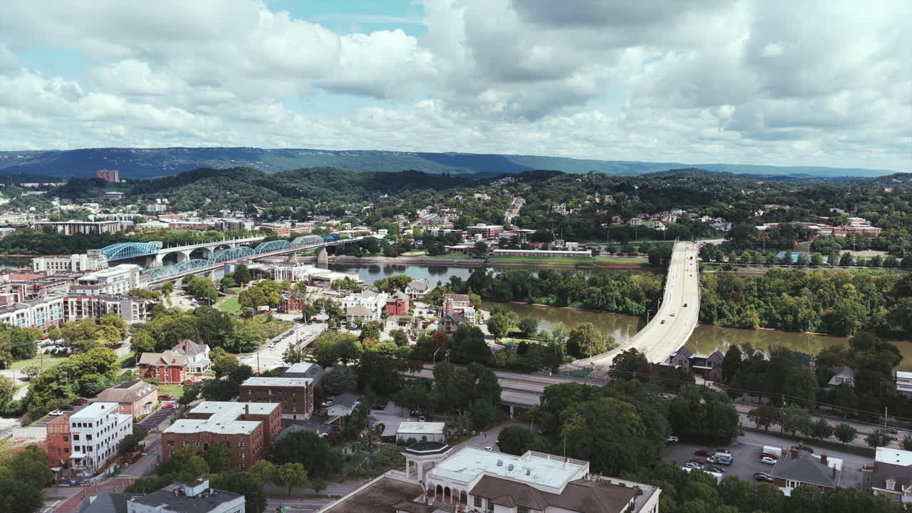 Drone footage captures Chattanooga’s bridges spanning the Tennessee River, framed by rolling green hills, city streets, and a bright summer sky