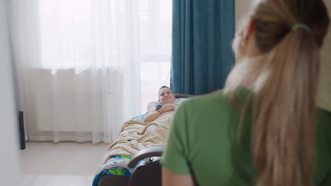 Young boy wakes up stretching in cozy bedroom covered with blanket smiling warmly as mother enters room to check on him sunlight shining through window curtain