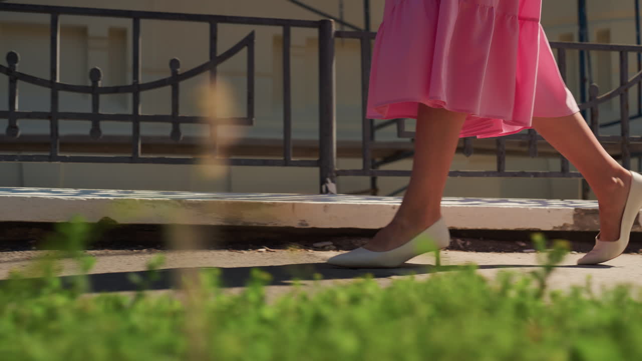 Donna in abito rosa che cammina lungo il marciapiede, a metà passo con tacchi bassi e gonna svolazzante con sfondo verde sfocato, ringhiera di metallo sullo sfondo, movimento dinamico e passo sicuro, luce solare calda, estate informale