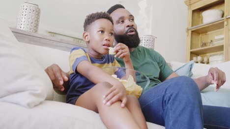 Glücklicher-Afroamerikanischer-Vater-Und-Sohn-Sitzen-Auf-Dem-Sofa,-Schauen-Fern-Und-Essen-Chips,-In-Zeitlupe