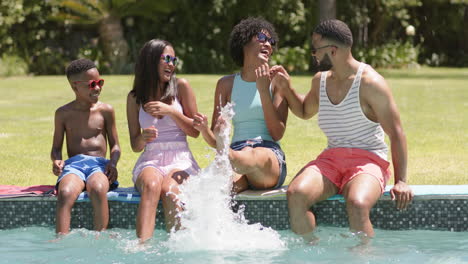 happy african american parents, son and daughter sitting in sun splashing, feet in pool, slow motion