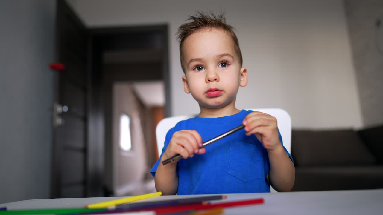 Encantador bebé niño con camiseta azul sentado en el escritorio sosteniendo un lápiz en las manos. El niño deja el lápiz y se levanta. Vista en ángulo bajo