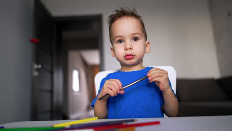 lovely baby boy in a blue t-shirt sits at desk holding a pencil in hands. child puts the pencil and stands up. low angle view.
