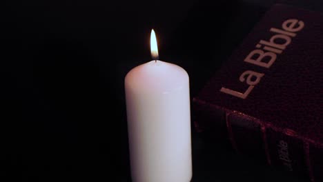 white holy candle flame burning next to bible against black background