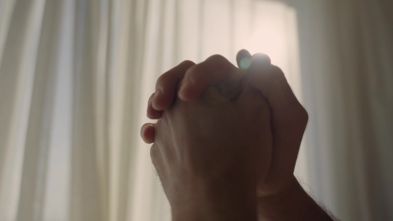 Silhouette delle mani di un uomo giunte in preghiera contro la luce brillante di una finestra