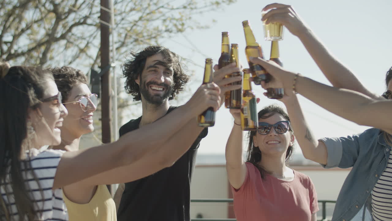 amigos felices tintineando botellas de cerveza mientras tienen una fiesta en la azotea en un día ventoso de verano