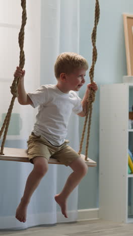 cheerful little boy in white t-shirt and shorts sits on wooden swing in nursery room with toys. male toddler laughs tilting head back in delight