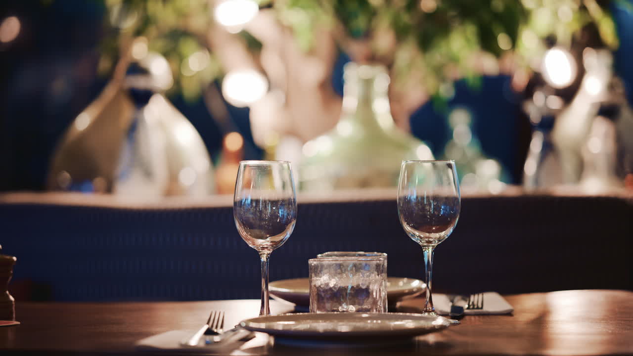 A neatly set dining table with two wine glasses, plates, and a candle, with decorative lights and bottles blurred behind