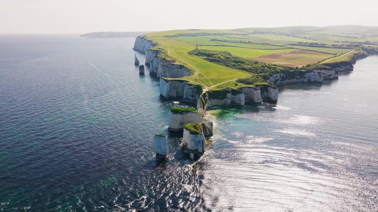 Laaja dronekuva ilmasta Old Harry Rocks -kallioista Dorsetin rannikolla Englannissa, esittäen dramaattisia valkoisia liitukivipylväitä, rannikkokallioita, luonnollisen merikaaren, vihreitä kallionharjanteiden polkuja ja viljelysmaata.