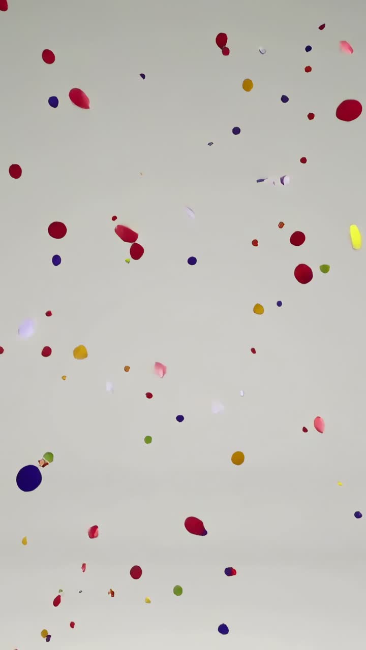 Video verticale: Pezzi di coriandoli colorati che cadono, volteggiando e ruotando verso il basso su uno sfondo grigio, scintillanti.