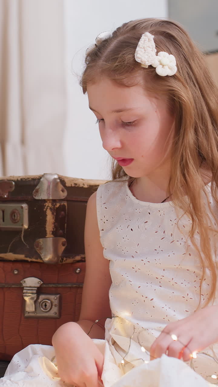 Caucasian girl examining tiny lights with concentrated expression, white dress, vintage trunk backdrop, hands working gently on wire frames portray curious tinkerer, patient craftsperson, and quiet