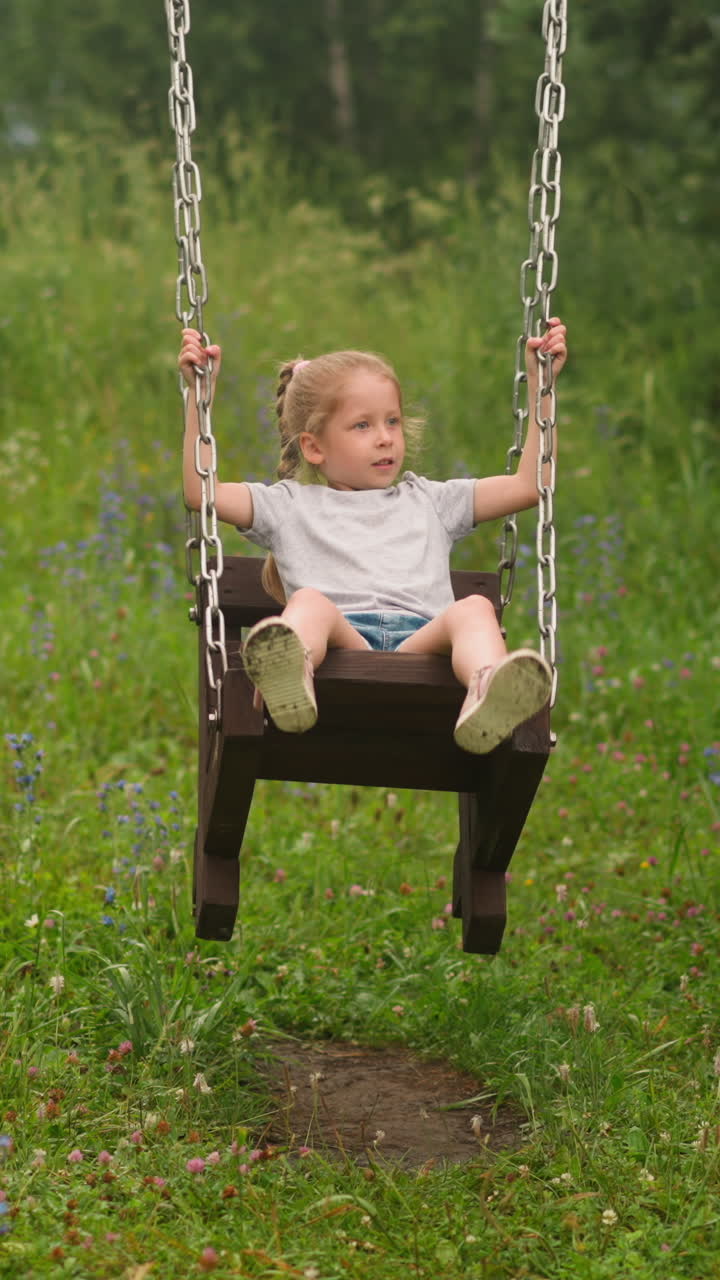 Pretty blonde little girl plays vintage wooden swings on ground against high meadow grass on overcast summer day at countryside slow motion