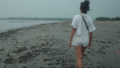 medium slow motion dolly shot of a young indian woman in white shirt and fanny pack while taking an evening walk on the beach during her summer vacation with a view on beautiful beach with calm sea