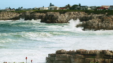 Waves-crash-into-rocky-coastline-with-houses-in-background-overlooking-sea